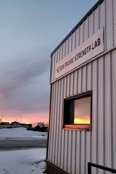 The sun sets behind Iron Prairie Strength Lab in Moosomin, SK, with a sign visible in the foreground.