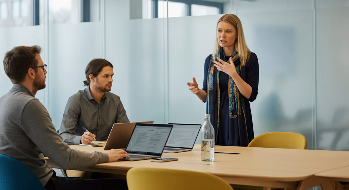 Drie volwassenen in een vergaderruimte, twee mannen zitten met laptops, een vrouw staat en spreekt toe.