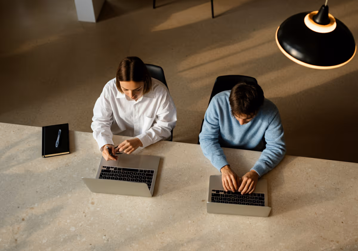 Twee mensen zitten naast elkaar aan een tafel en gebruiken laptops in een goed verlichte werkruimte.