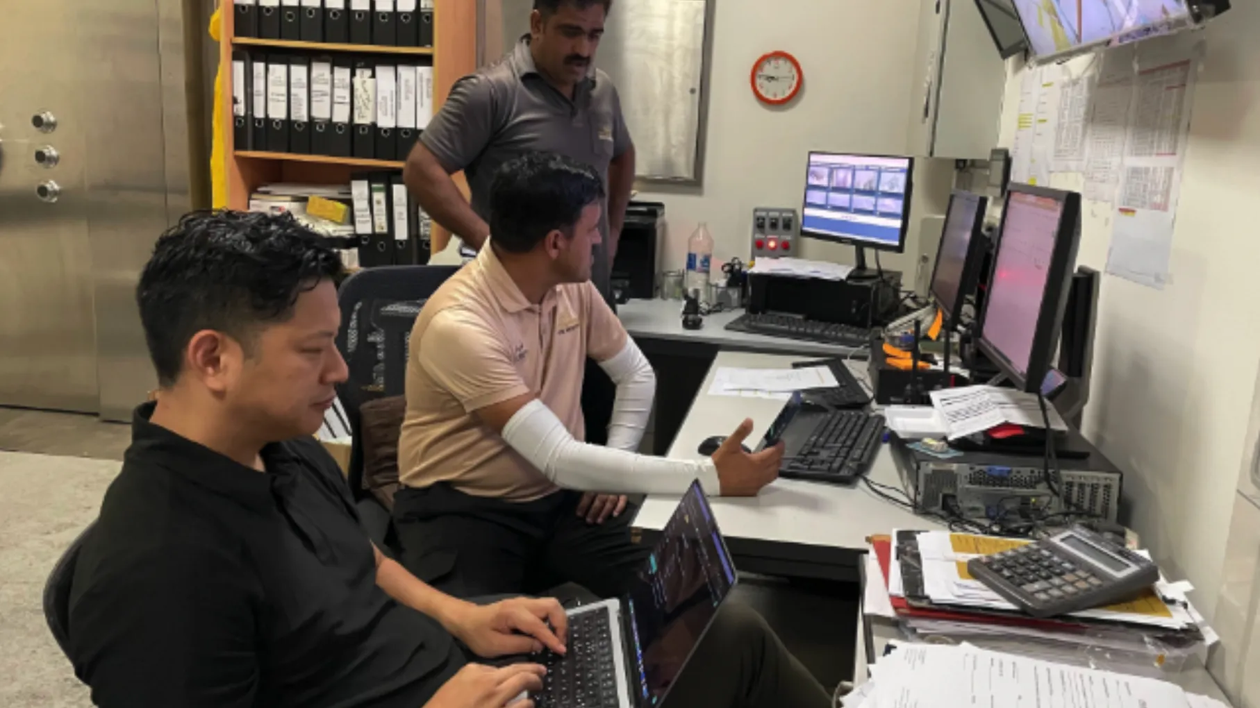 Three men working in a control room with multiple computer monitors and files, one using a laptop, another holding a smartphone, and the third standing behind.