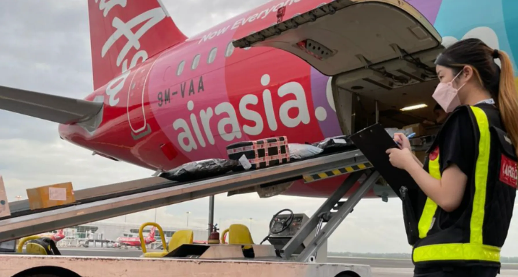 Woman in safety vest and face mask checking cargo on clipboard beside AirAsia plane being loaded with parcels.