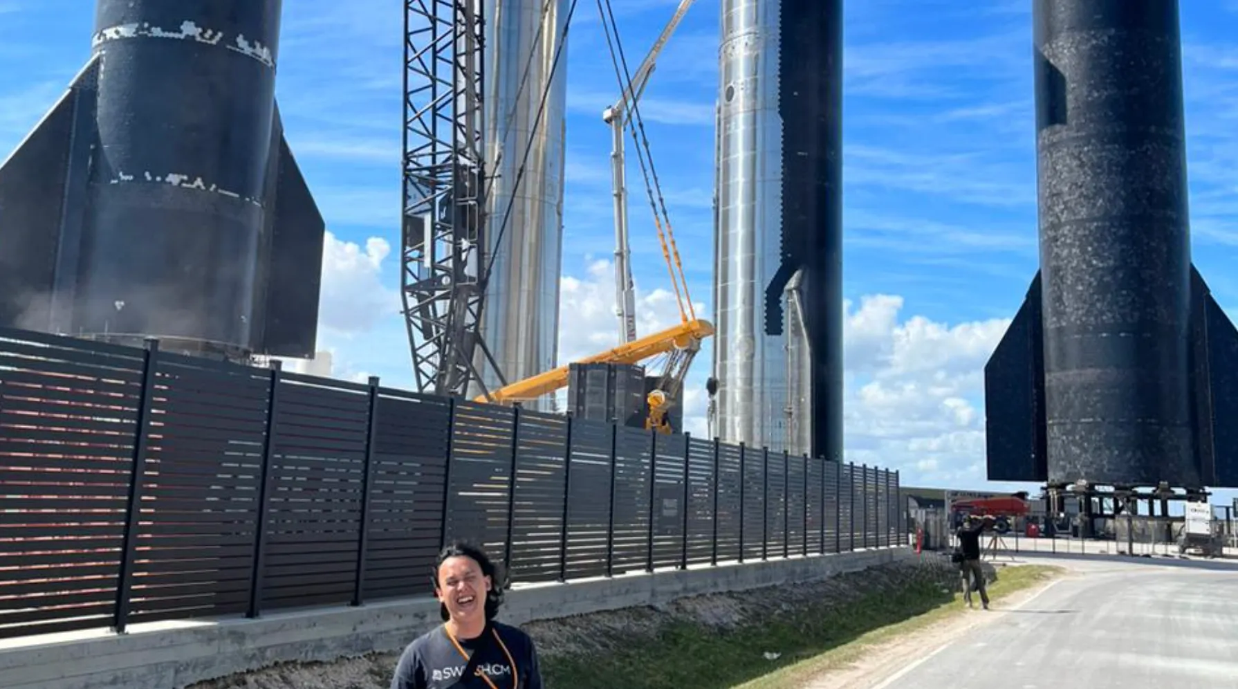 Person smiling in front of a fence with large rocket prototypes and cranes at a construction site under a blue sky.