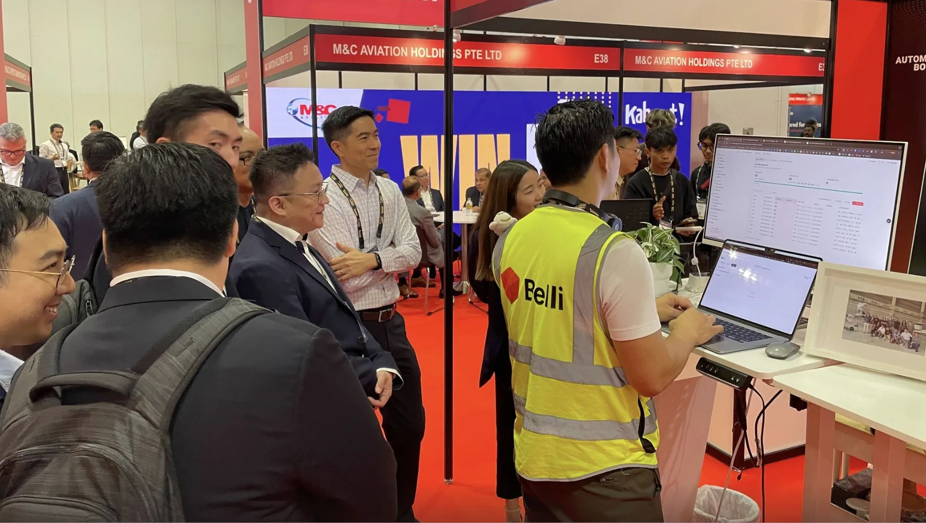 Group of people interacting at an exhibition booth with a man in a yellow Belli vest working on a laptop and a large monitor displaying data.