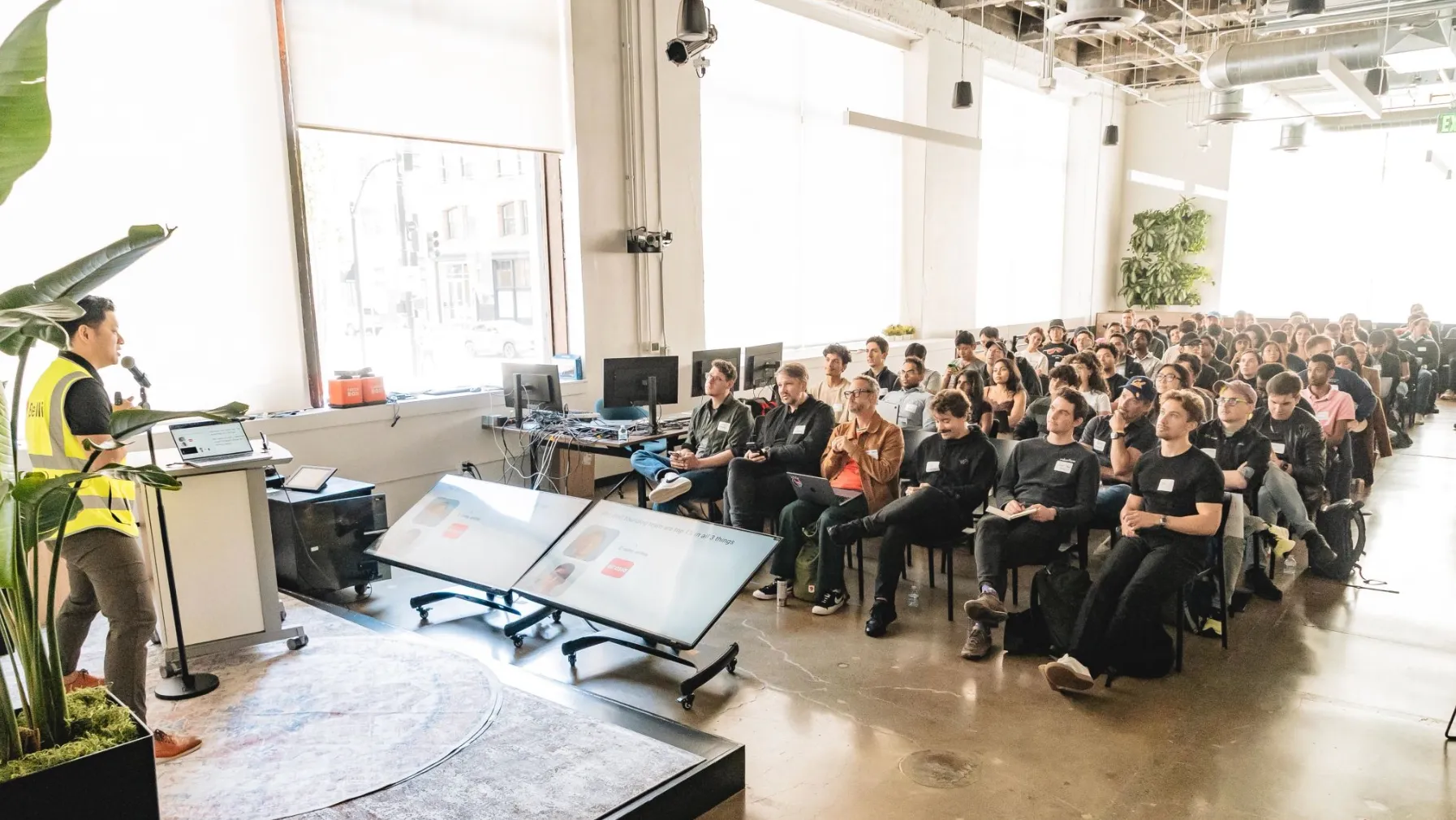 Speaker in a yellow vest presenting with a microphone to a seated, attentive audience in a bright, modern conference room.