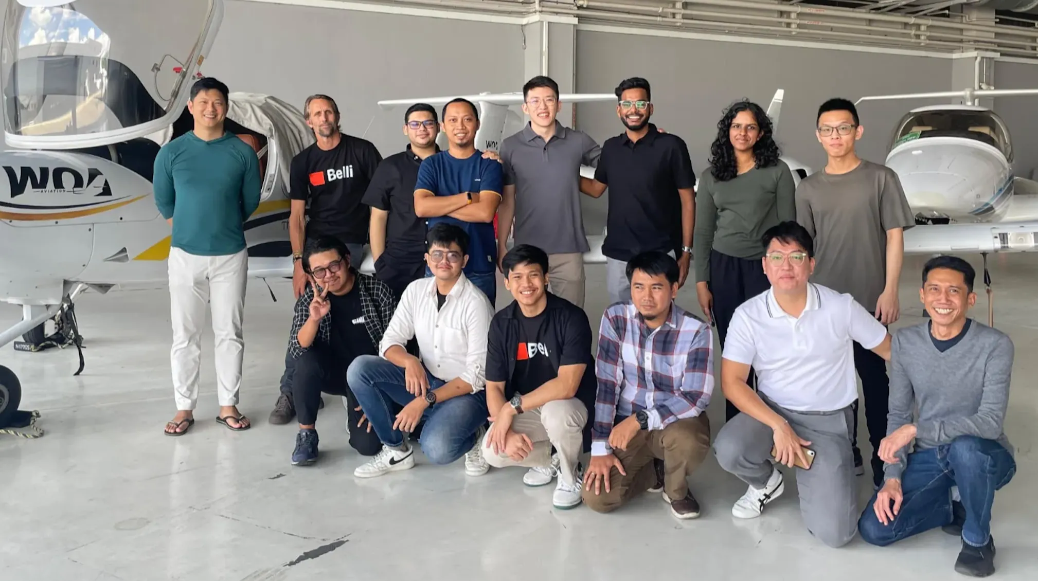 Group of 14 people posing together in an aircraft hangar with two small planes in the background.