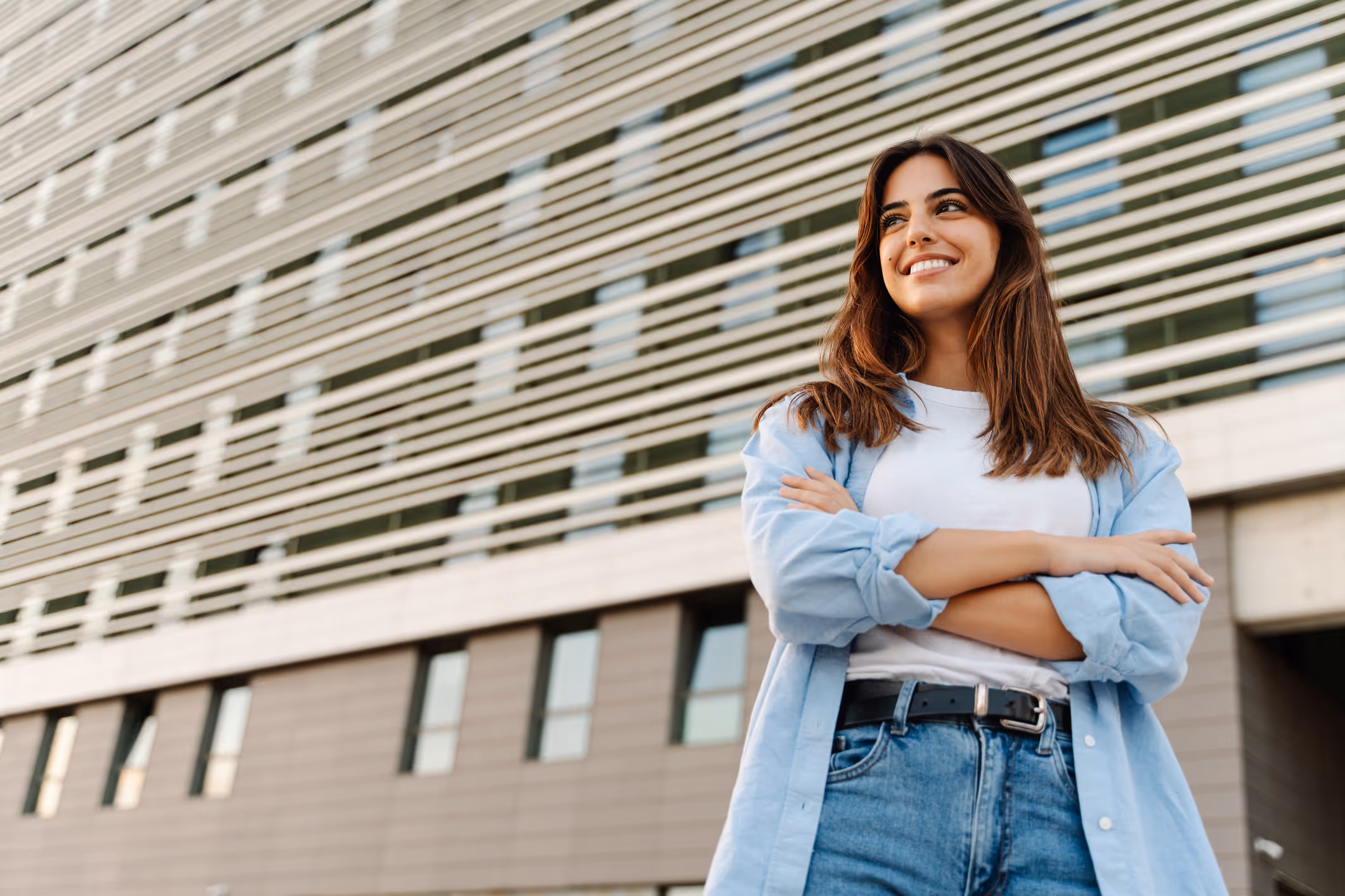 Confident young woman with brown hair standing with arms crossed in front of modern building.