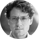 Black and white close-up portrait of a man with curly hair, glasses, and a serious expression.