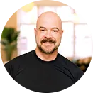 Smiling bald man with a beard wearing a black shirt, sitting indoors with blurred windows and plants in the background.