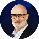 Portrait of a bald man with a gray beard and glasses wearing a white shirt and black blazer against a dark blue background.