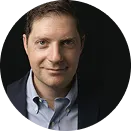 Portrait of a man with short brown hair wearing a dark blazer and light blue shirt against a black background.