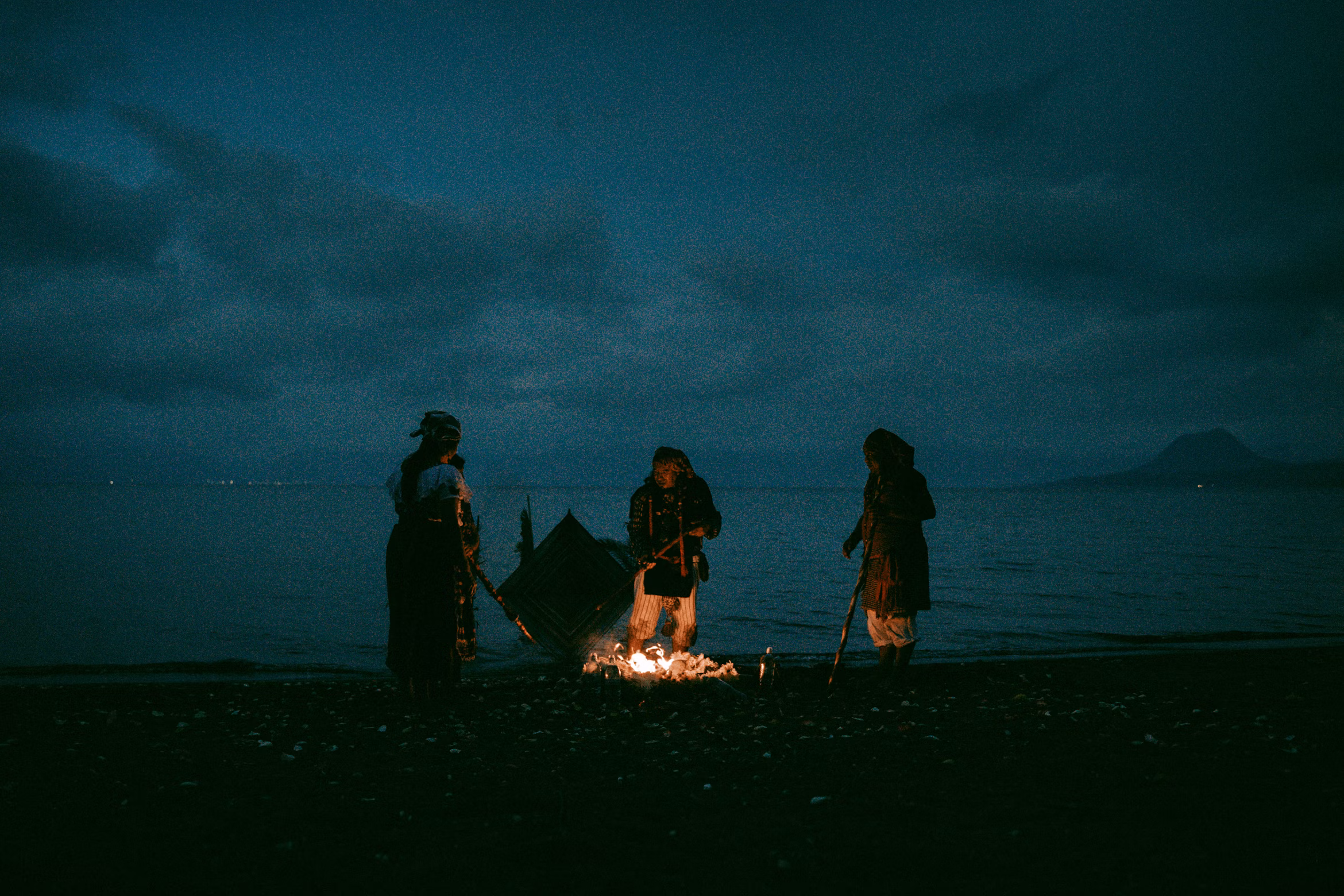 Three people gathered around a fire by the sea at night, ritual or ceremony scene in atmospheric photography
