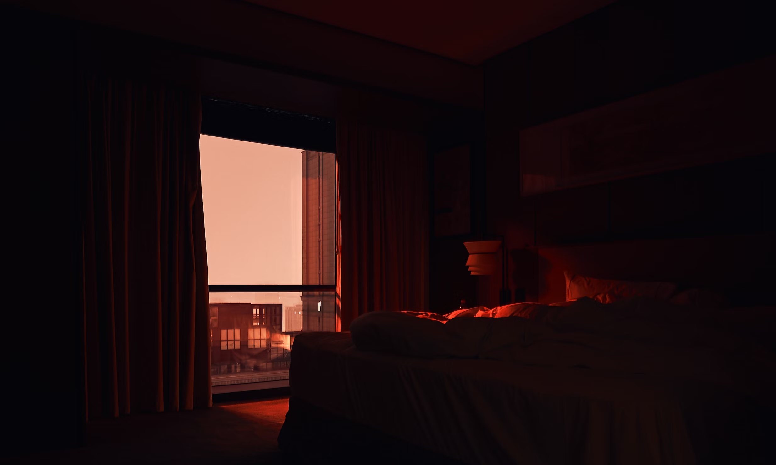 Moody dimly lit bedroom with unmade bed facing a large window showing city buildings at sunset glow.