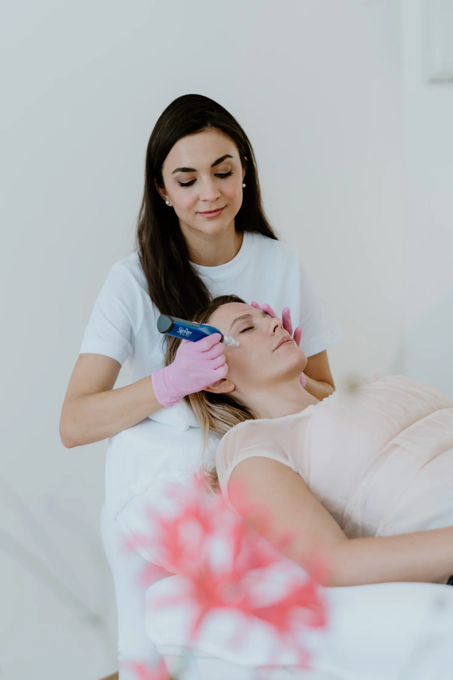 Eine junge, dunkelhaarige Frau in weißer Kleidung behandelt das Gesicht einer liegenden Frau mit einer Microneedling-Pen.