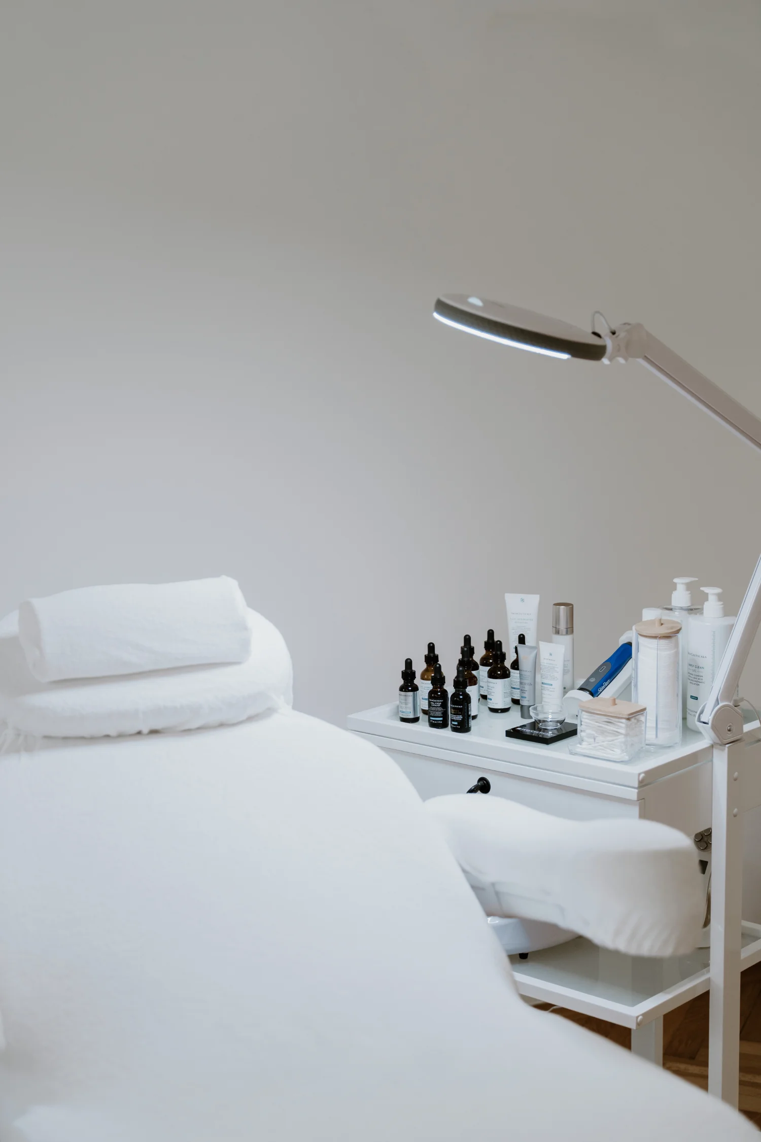 Eine weiße Behandlungsliege, daneben ein weißes Tischchen, auf welchem Pflegewirkstoffe und eine Microneedling-Pen stehen.