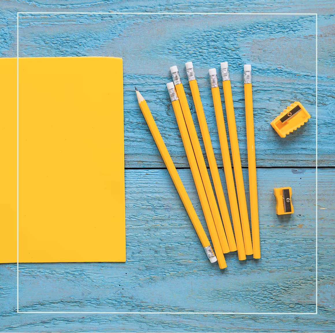 "Yellow notebook with sharpened pencils and pencil sharpeners on a bright blue wooden desk, flat lay."