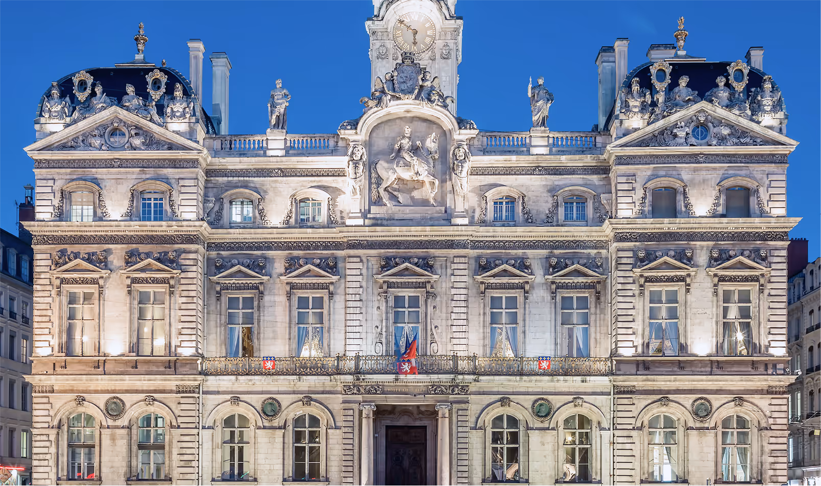 “Illuminated baroque-style façade of a grand historic building at night, with statues, clocktower, and ornate stone carvings.”