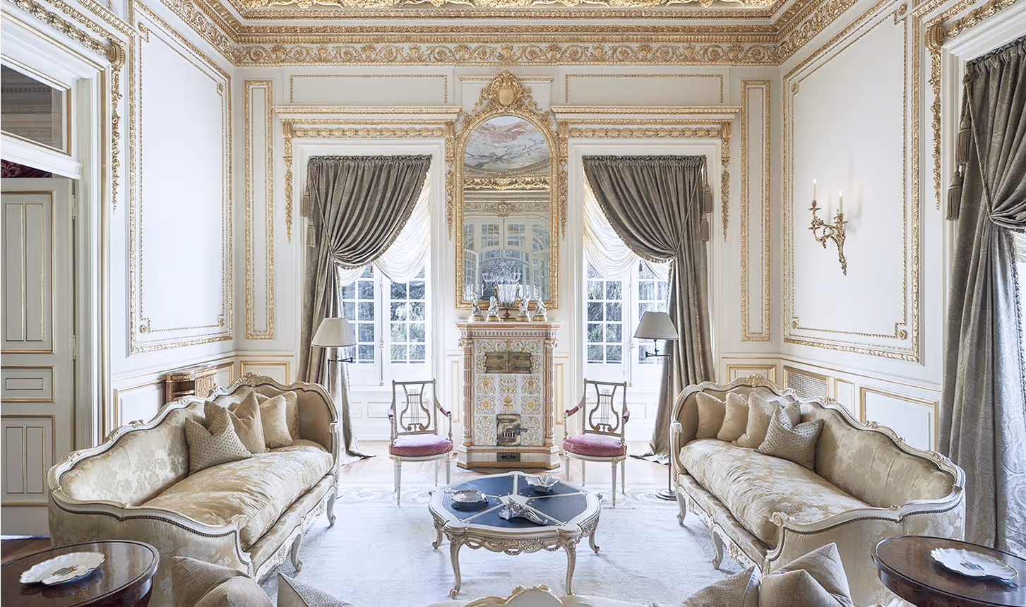 “Elegant drawing room with gilded plasterwork, tall sash windows, silk drapes, and antique sofas around a decorative fireplace.”