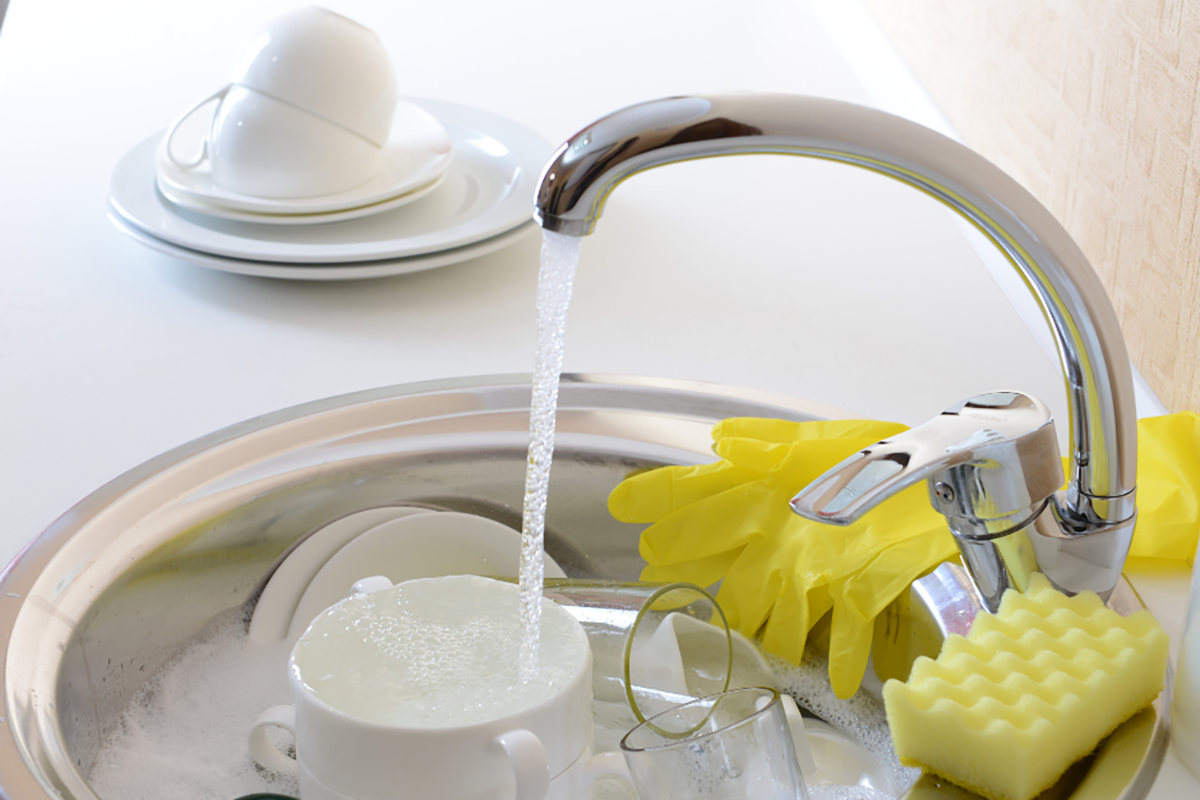 A running kitchen sink tap; the sink is filled with cups and glasses to be washed.