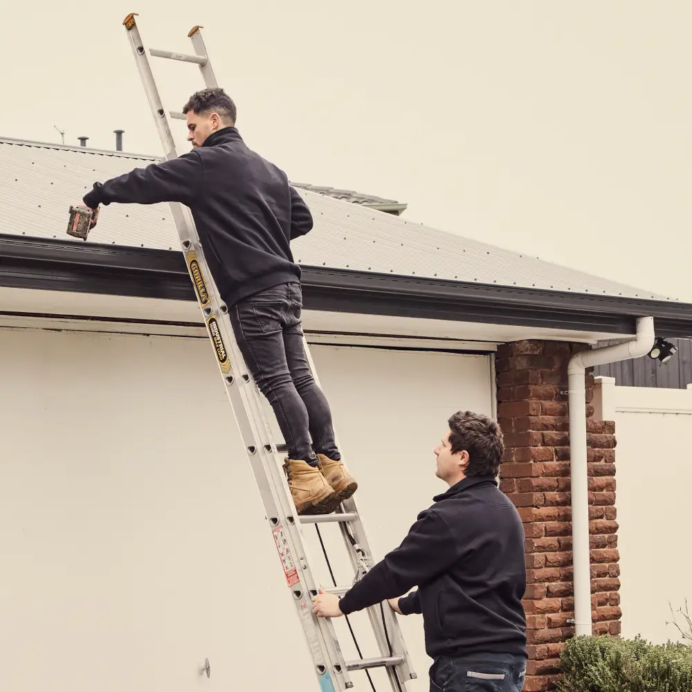 Two workers on ladder repairing roof gutters near brick wall