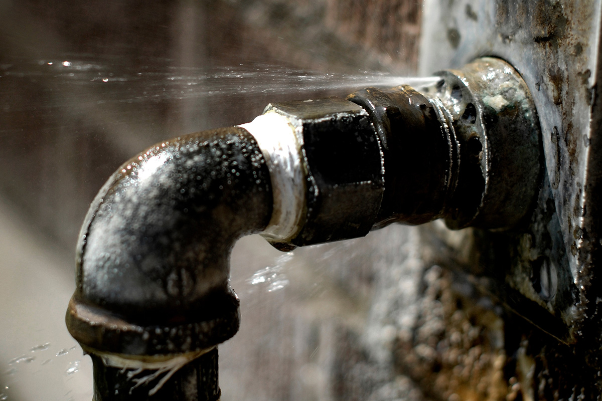 An old metal pipe with pressurised water squirting through a tiny crack in a join. Waterproof silicone sealant can be a temporary fix for a leaking pipe like this, but it’s not a long-term fix.