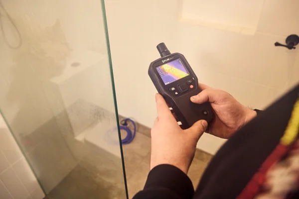 Plumber using a thermal imaging camera to detect water leaks inside a shower area.
