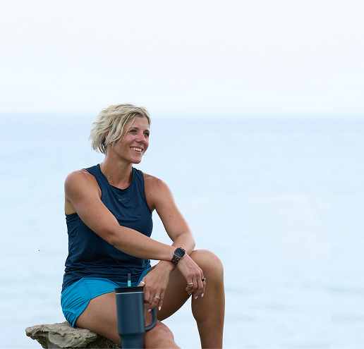 Ruth sitting on a rock and holding a drinking flask, with the sea in the background.
