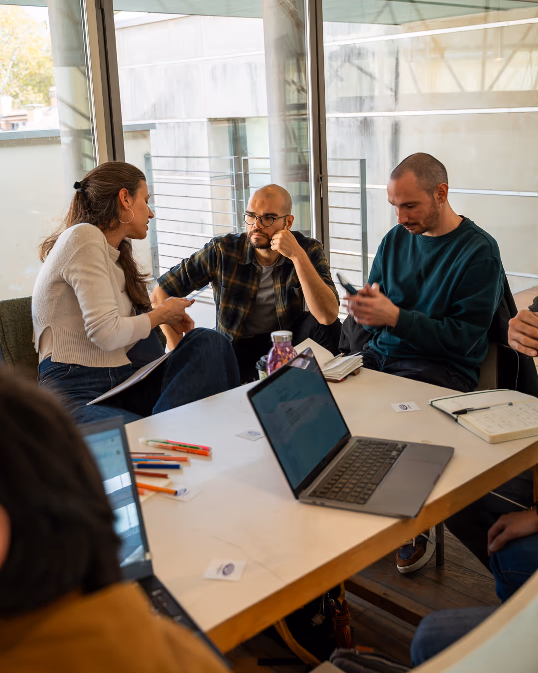 Quattro persone sedute intorno a un tavolo con laptop, quaderni e penne, coinvolte in una discussione in un ufficio moderno con grandi finestre.