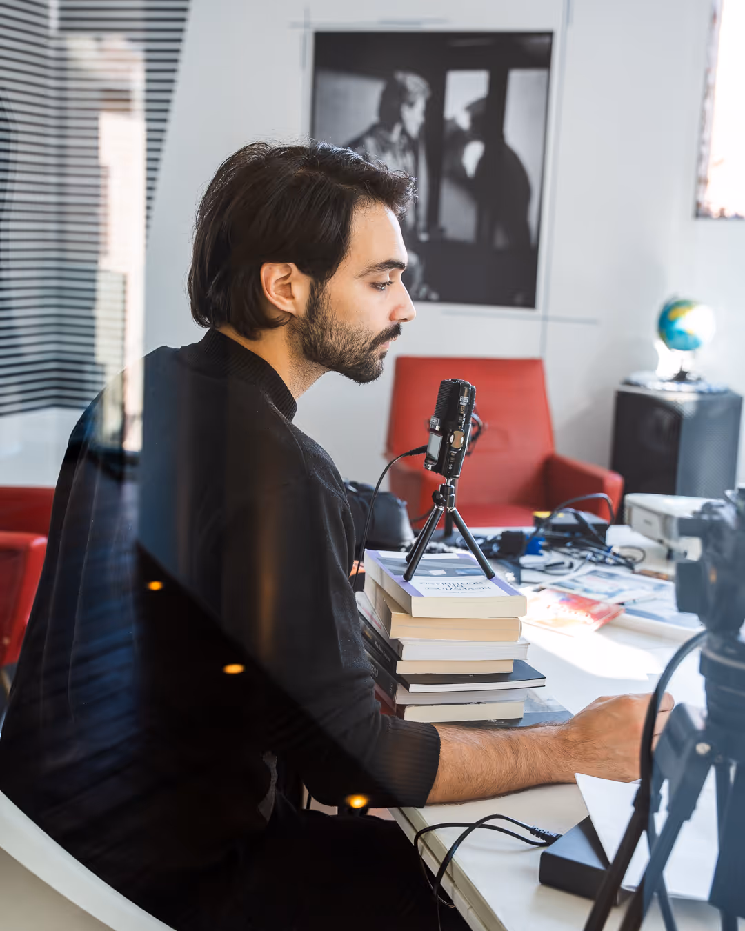 Uomo seduto a una scrivania con microfono su un supporto posizionato su una pila di libri, che utilizza un computer in un ambiente di lavoro con poltrona rossa e mappamondo.