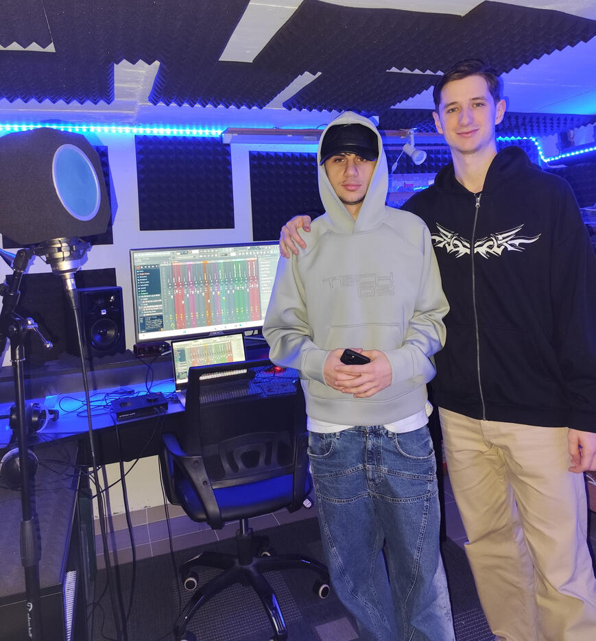 Due giovani uomini posano in uno studio di registrazione musicale. Uno indossa una felpa grigia con cappuccio e cappellino nero, l’altro una felpa nera con zip e pantaloni chiari. Sullo sfondo si vedono due monitor con un software di produzione musicale aperto, casse da studio, microfono con filtro anti-pop e illuminazione LED blu.