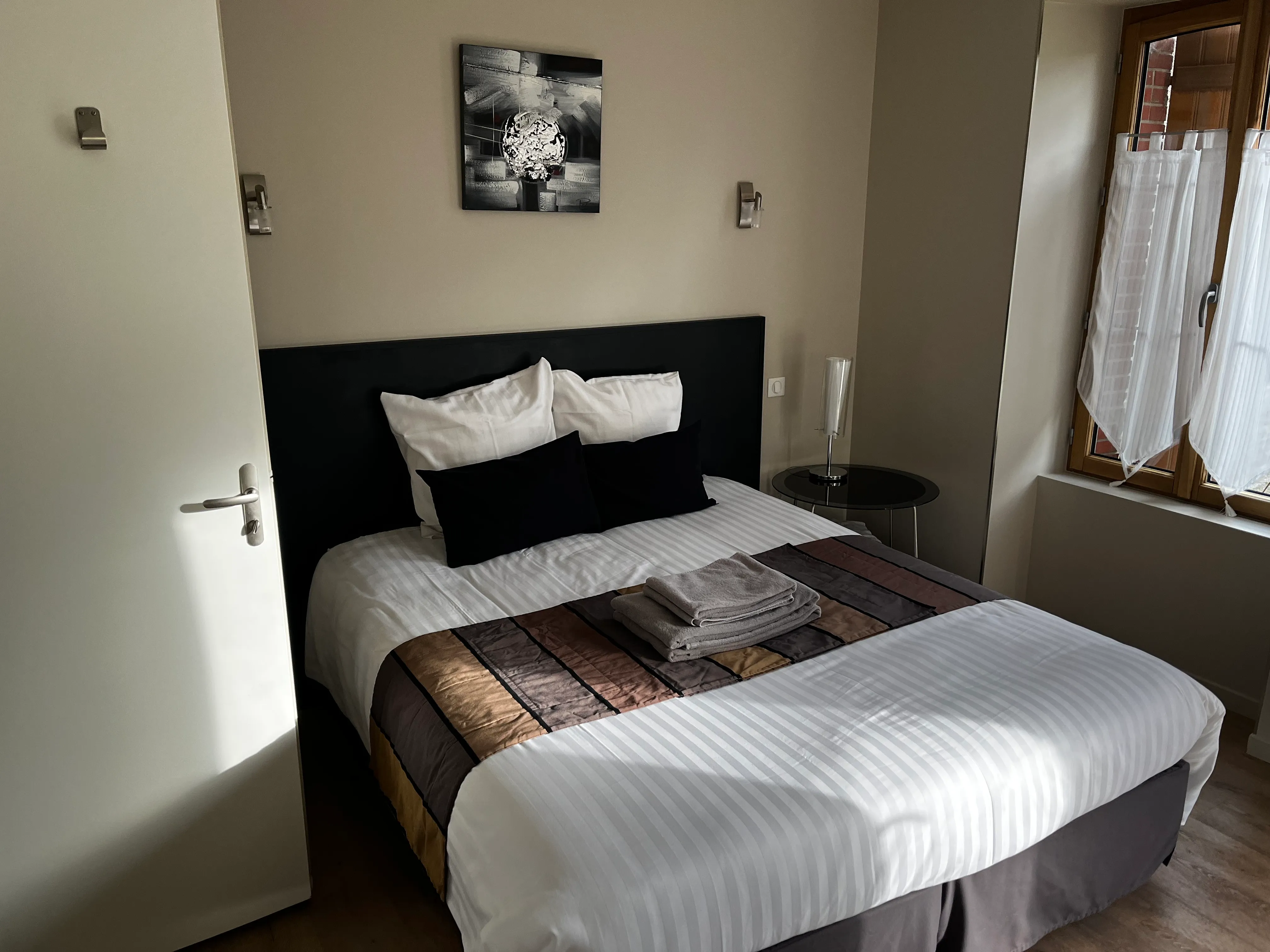 Chambre avec lit double, linge de lit blanc, coussins noirs et blancs, couverture colorée, serviettes pliées et fenêtre avec rideaux blancs.