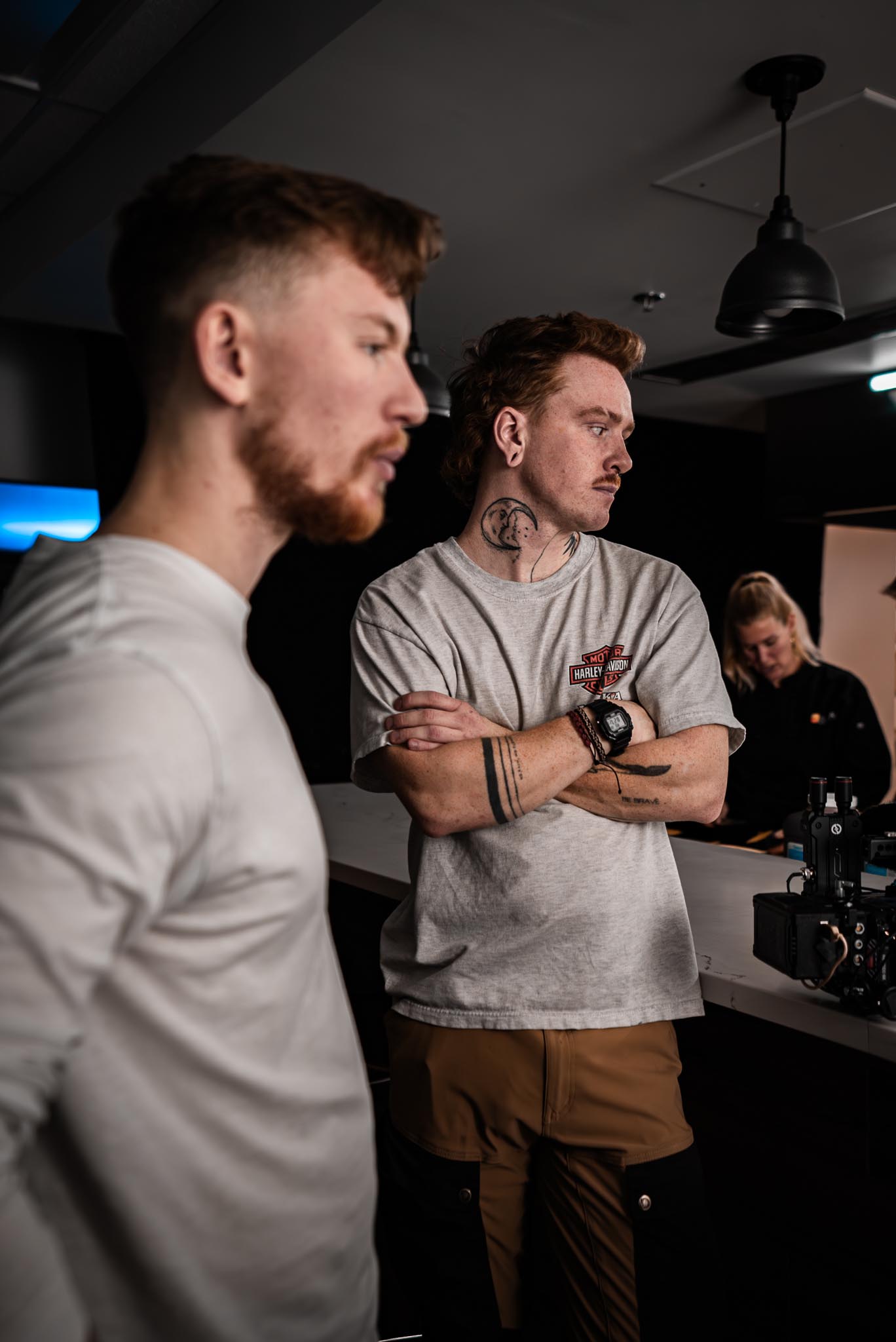 Two men in a dimly lit room, one with tattoos and a watch crossing his arms, a woman in the background working at a counter.