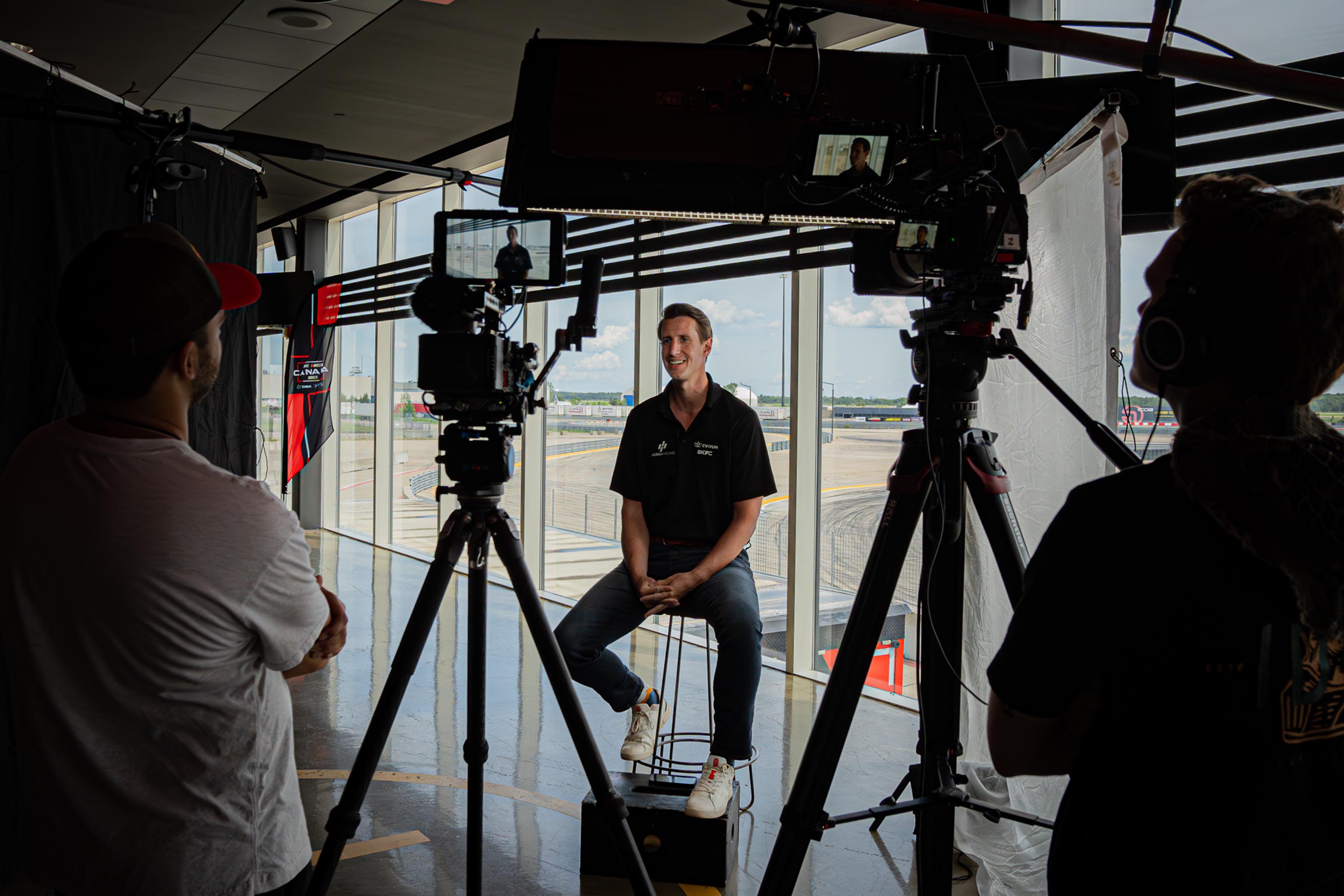 Man in a black shirt sitting on a stool in front of large windows, being filmed by two cameramen with professional video cameras indoors.