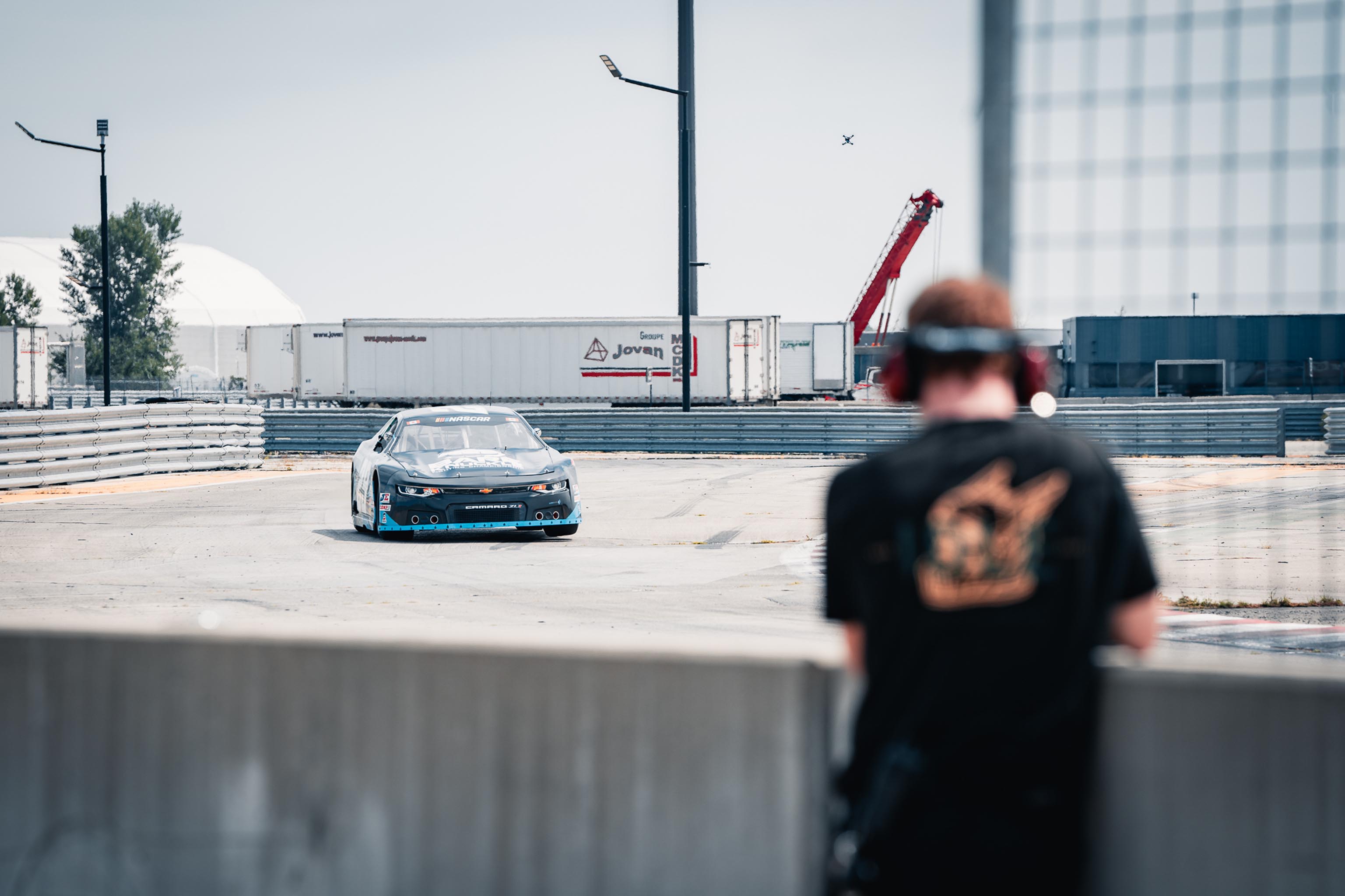 Voiture de course NASCAR Chevrolet Camaro ZL1 bleue sur une piste avec un homme flou en avant-plan portant un casque antibruit.