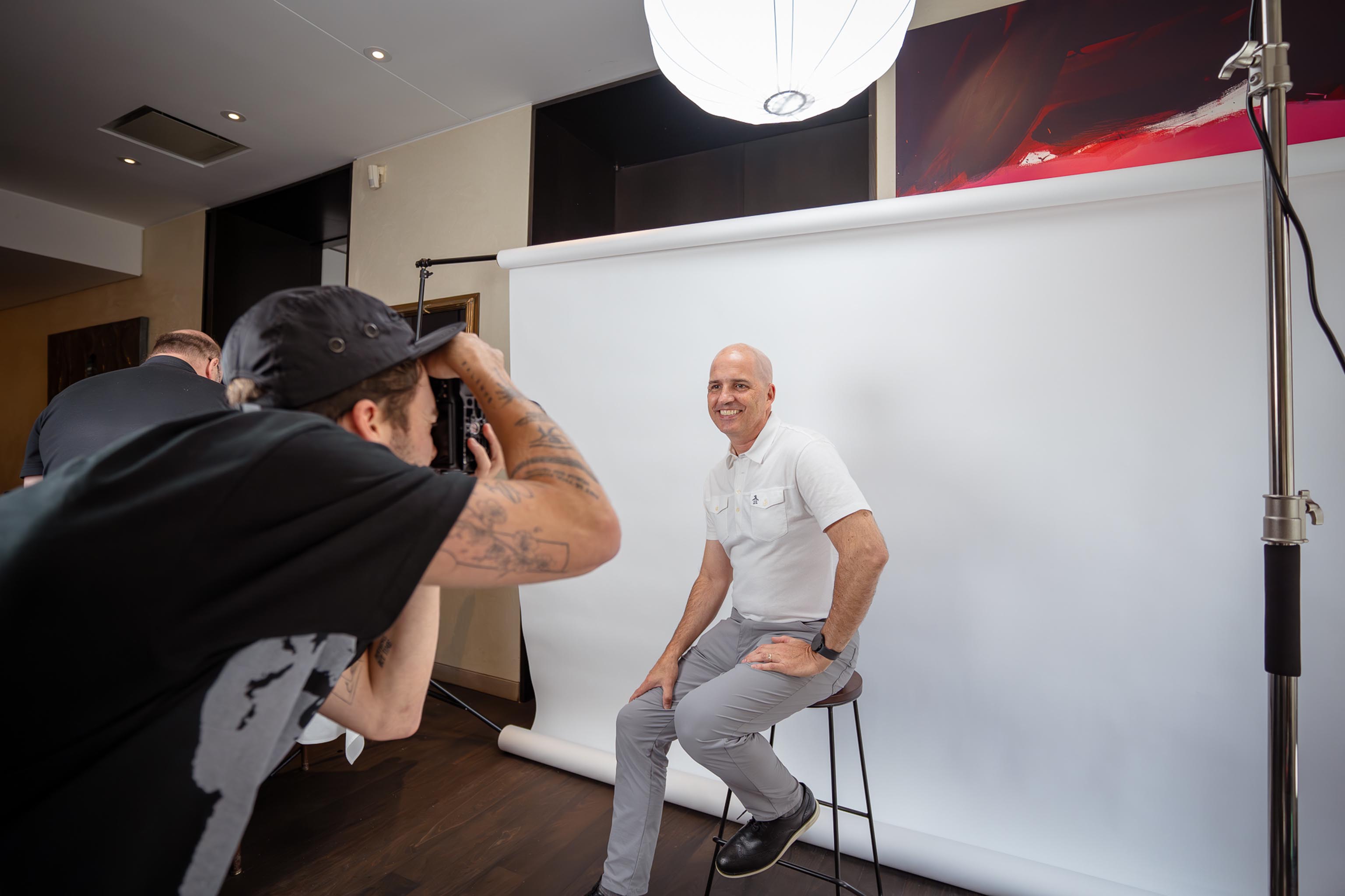 Un photographe tatoué prend une photo d'un homme souriant assis sur un tabouret devant un fond blanc en studio.