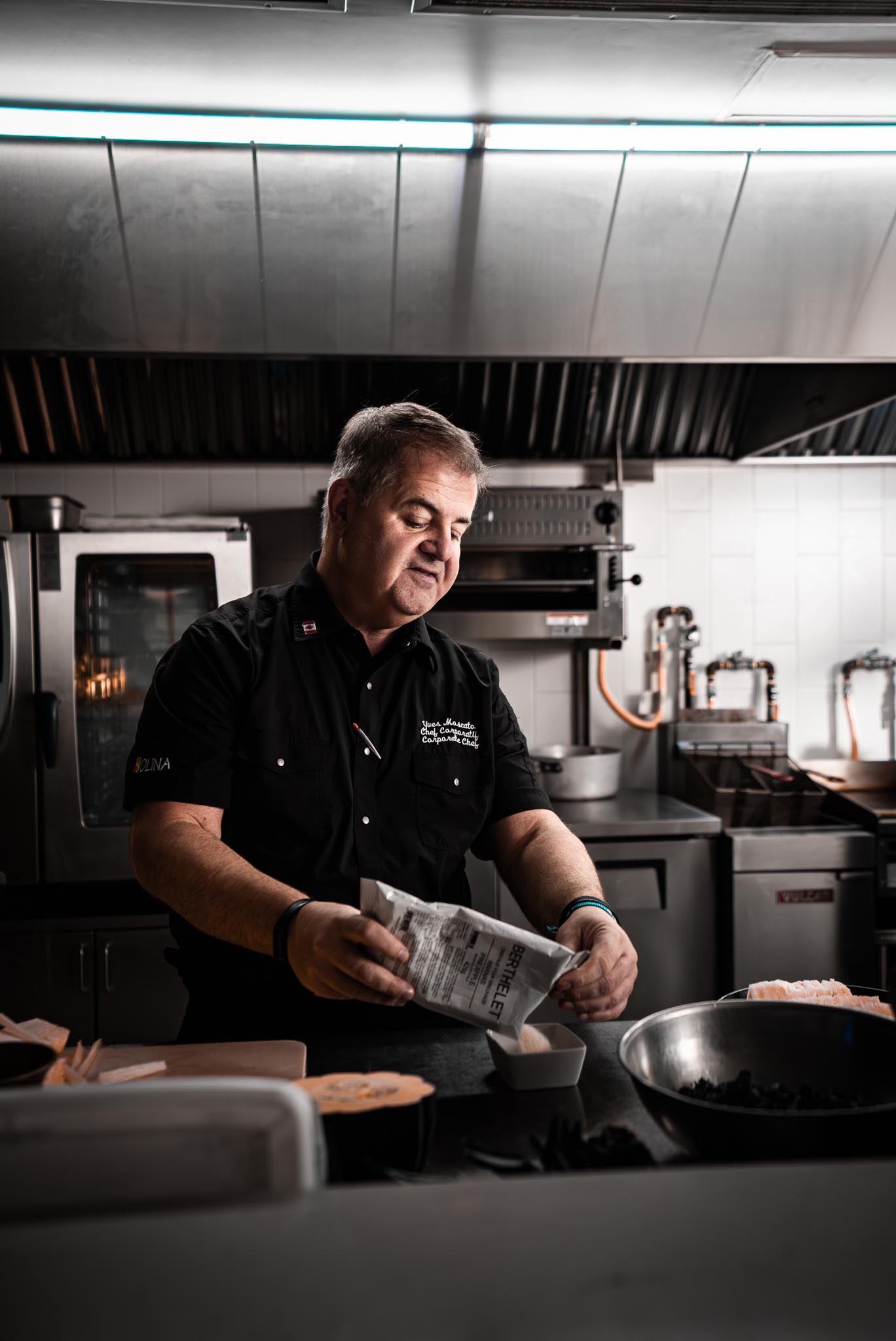 Chef en chemise noire dans une cuisine professionnelle, versant un ingrédient d'un paquet dans un bol blanc.