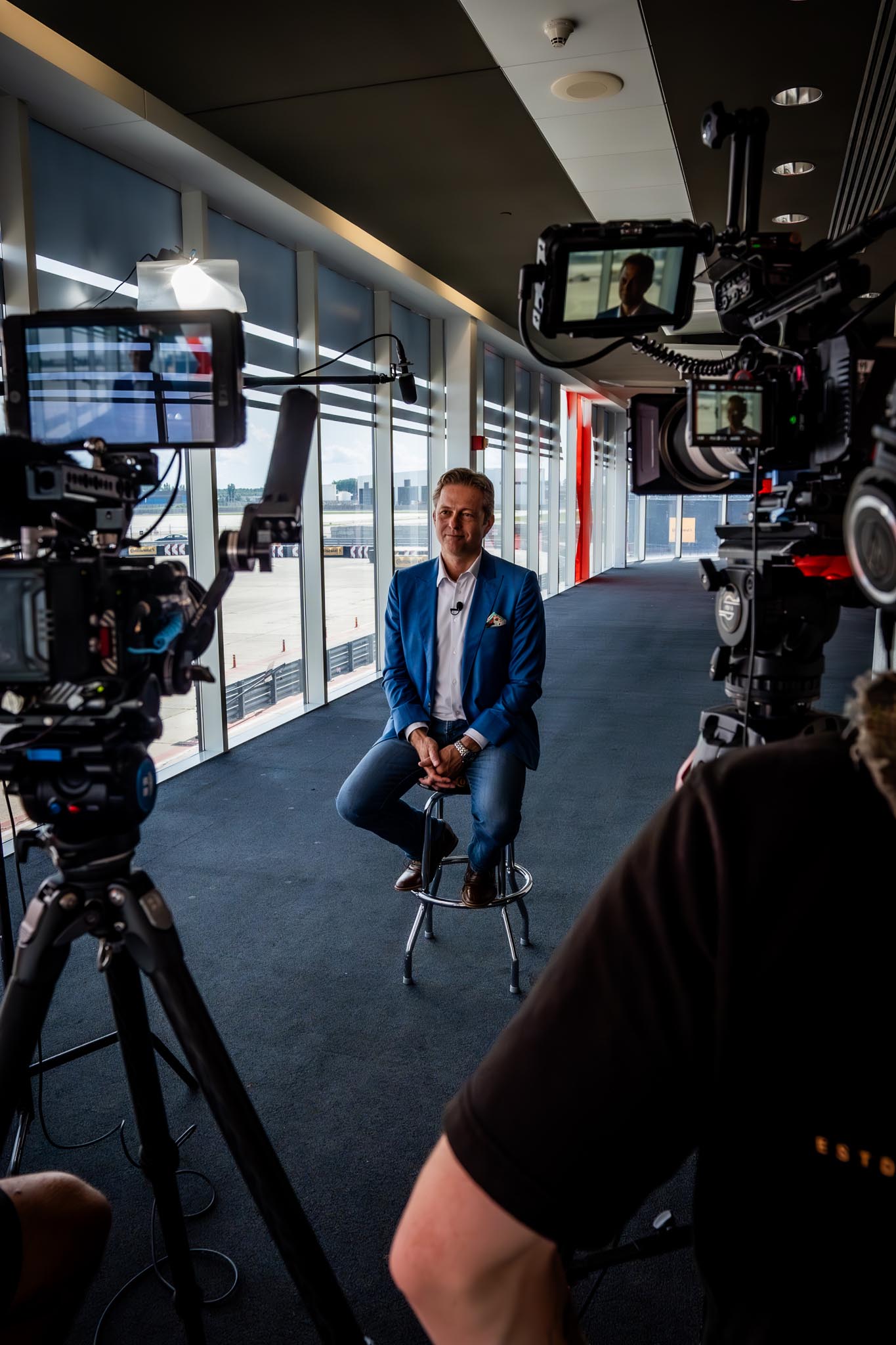 Un homme en veste bleue assis sur un tabouret dans un couloir vitré, filmé par plusieurs caméras professionnelles.