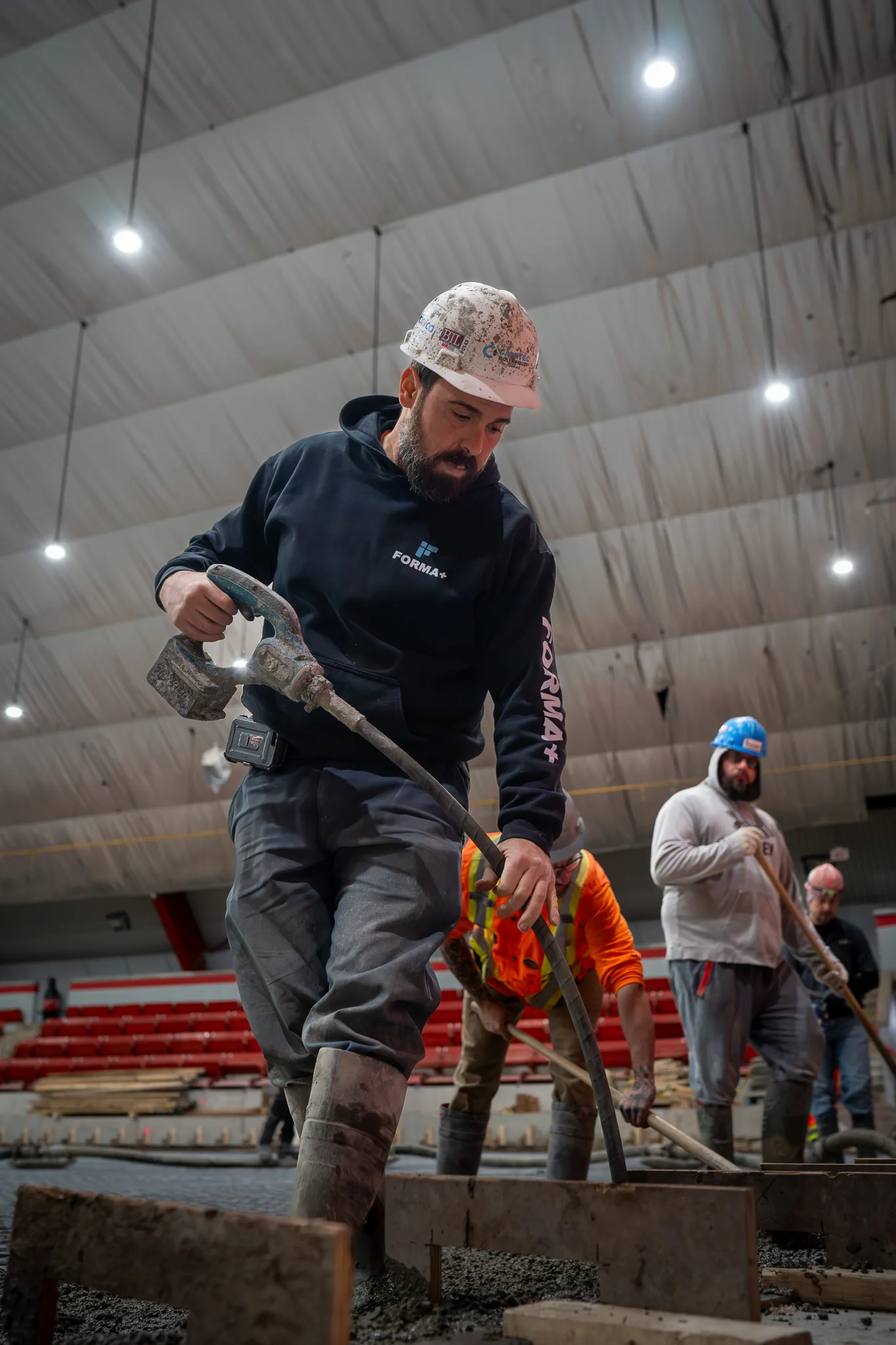 Construction worker wearing a hard hat and black hoodie using a vibrating tool on wet concrete indoors with other workers smoothing the surface.