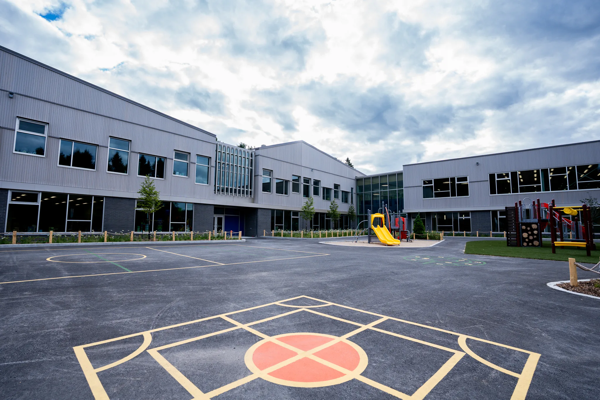 Empty school playground with painted game markings, two yellow slides, and modern school building under a cloudy sky.