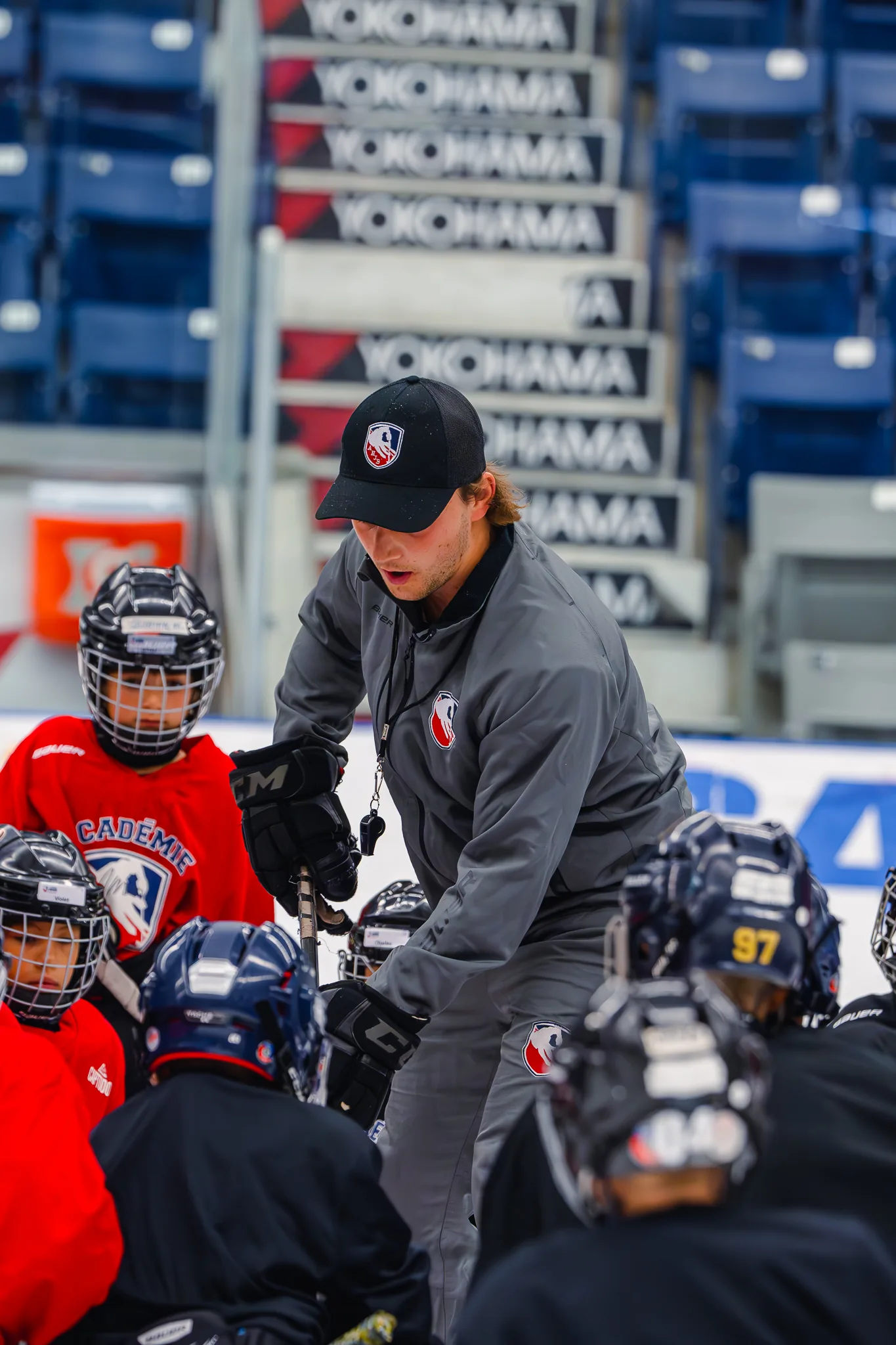 Hockey coach in gray jacket and black cap instructing young players on ice rink.