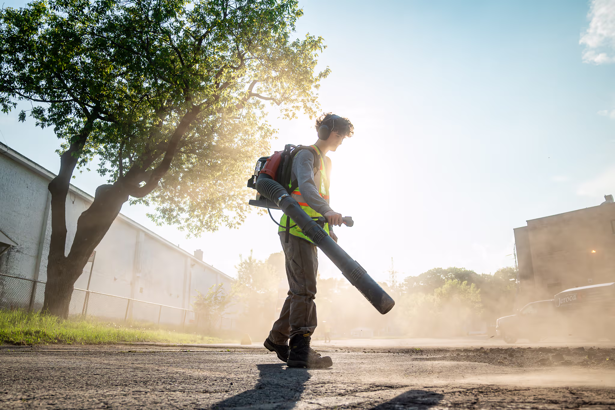 Jeune homme portant un gilet de sécurité et un casque anti-bruit utilise un souffleur de feuilles dans une zone urbaine ensoleillée avec de la poussière en suspension.