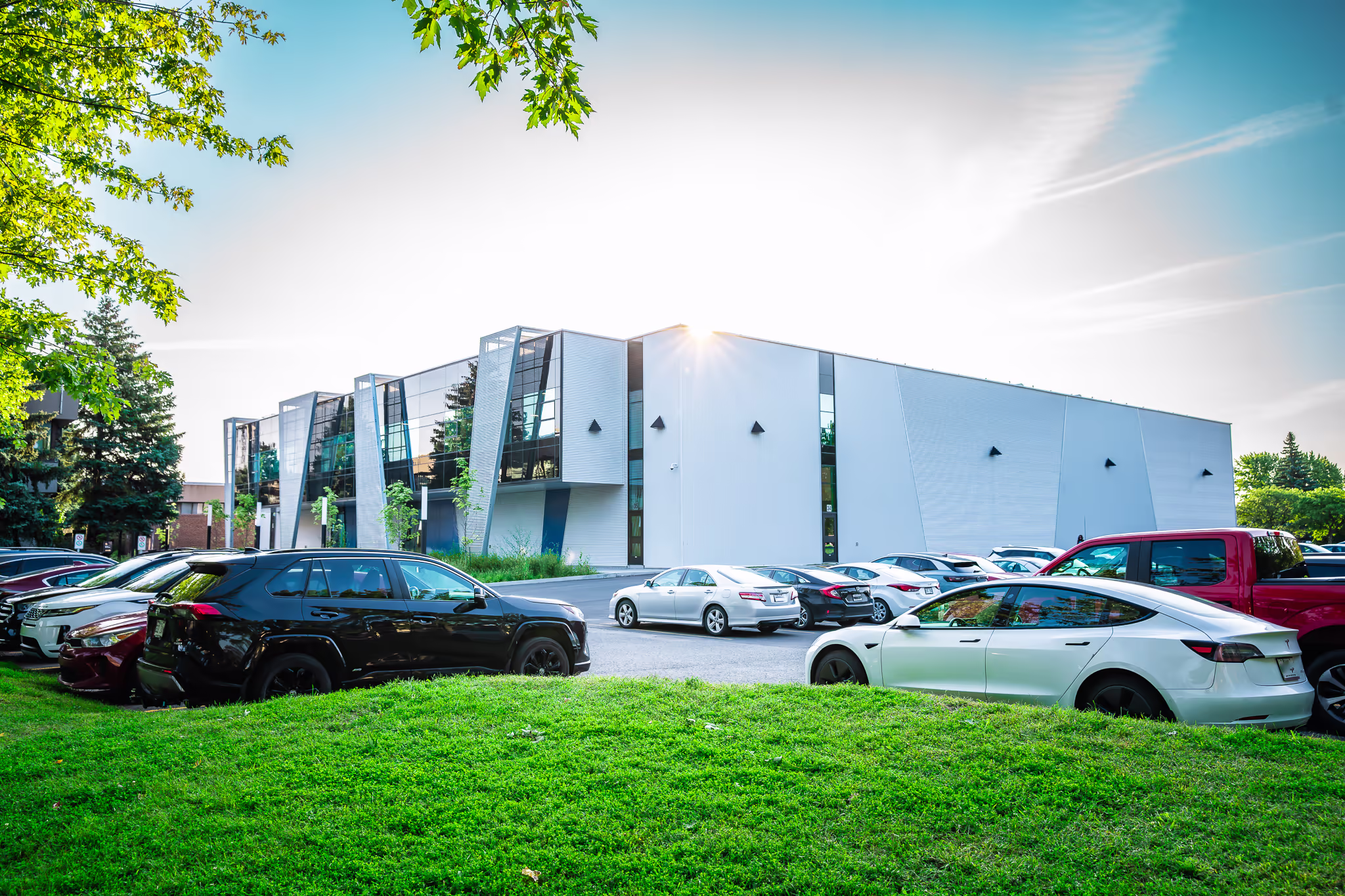 Édifice moderne à deux étages avec grande façade en verre et blanc, espace de stationnement extérieur avec plusieurs voitures garées, pelouse verte en avant-plan.