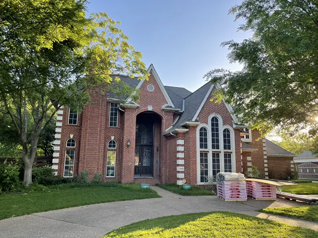 Two-story red brick home with new roofing materials stacked in front, prepared for installation by CLC Roofing.