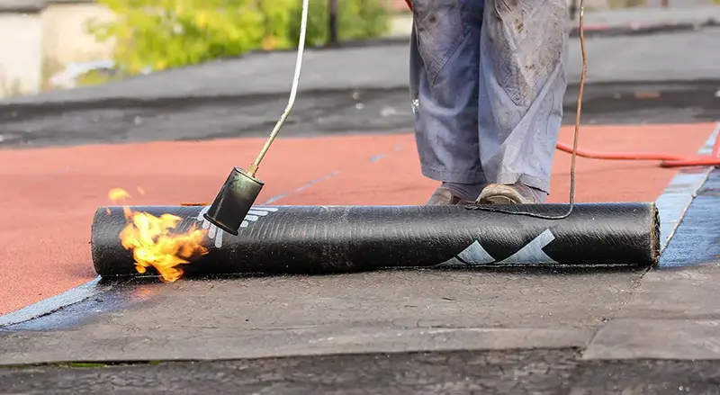 Roofer applying torch-down roofing membrane during flat roof installation on a commercial building.
