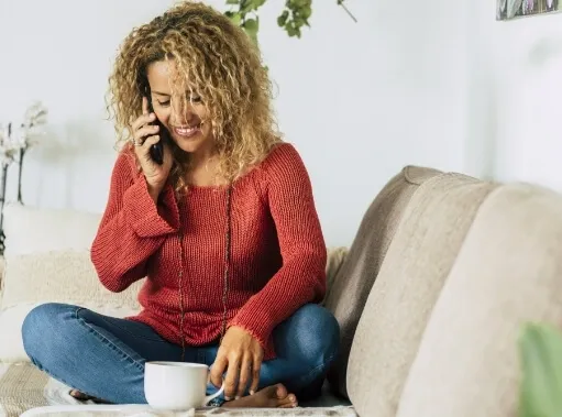 A woman with curly hair sits on a couch, smiling while talking on the phone and enjoying a cup of coffee.