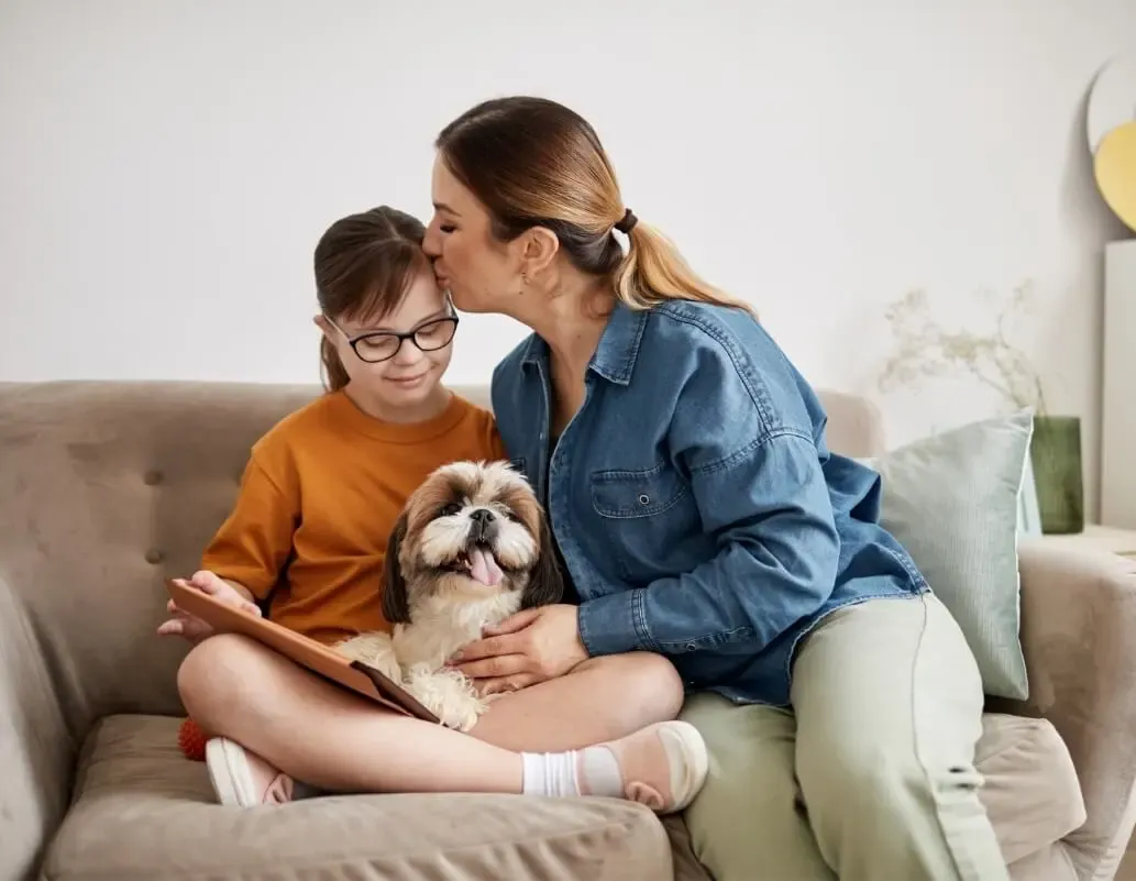 A mother kisses her daughter while they relax with a dog on a cozy couch.
