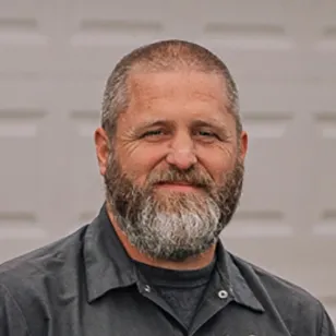 A man with a gray beard and shirt poses confidently outside a garage.