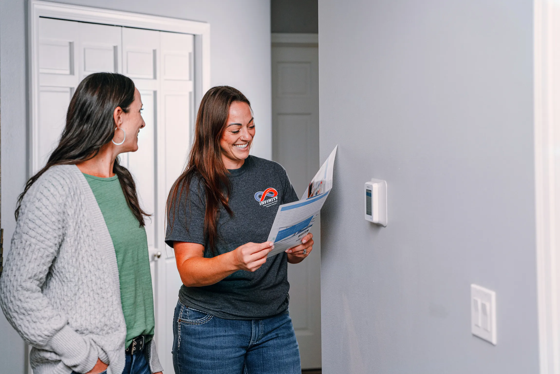 Two women discuss a thermostat while reviewing an informative brochure in a well-lit hallway.

