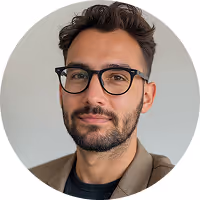 Man with short dark hair and beard wearing black glasses and a brown jacket, looking at the camera against a light gray background.