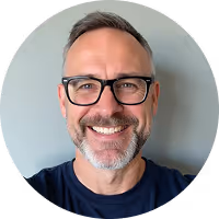 Smiling middle-aged man with glasses, gray hair, and a beard wearing a dark shirt against a plain background.
