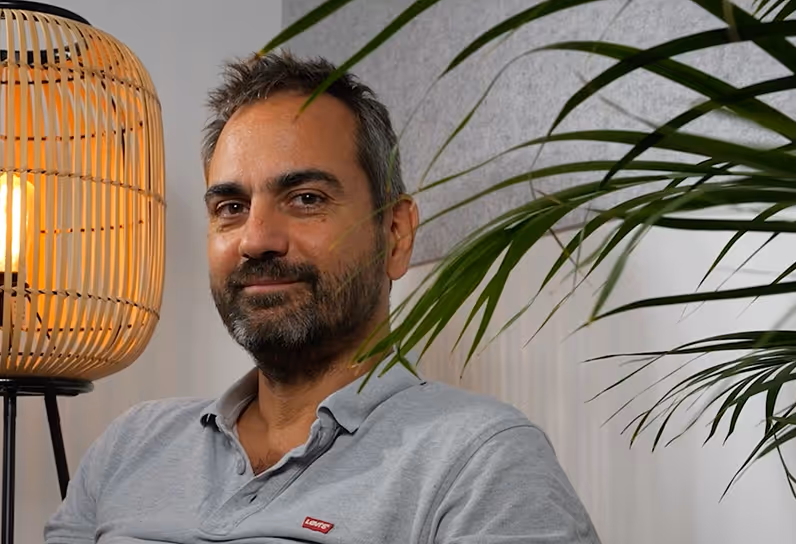 Man with short dark hair and beard wearing a gray Levi's polo shirt sitting near a wicker lamp and green indoor plants.